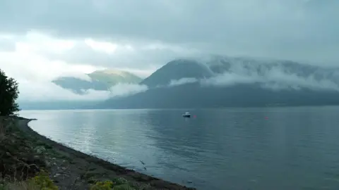 A stunning landscape of a Scottish loch, calm and empty, save a lone small fishing boat. Mountains frame the background and mist weaves its way up and down the outline of the hills