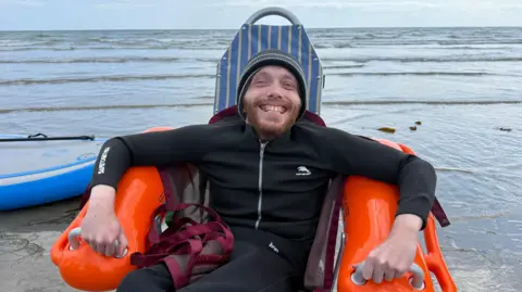 Jordan McGarel in a floating chair with orange arms and blue back. He is wearing a black wetsuit, a red life jacket, and a grey hat. He is at the water's edge.