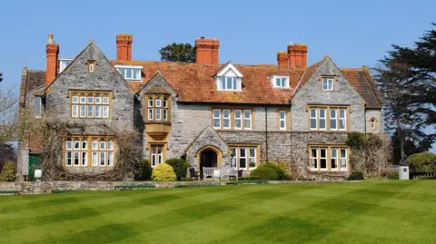 A large historic looking boarding school with grey pointed bricks, terracotta tiled roof and lots of bay windows. There is a bright green manicured lawn in the foreground, and various climbing plants on the side of the building. 