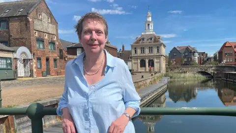 Alison Gifford stands on the King's Lynn Quayside with water behind her. On the quay is a small square stone building which is the Custom House. Alison is wearing a blue shirt and a string of white pearls. She has short grey hair and is smiling at the camera. 