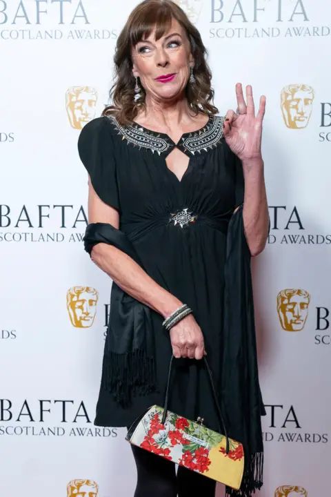 PA Media Doon MacKichan, wearing a short black dress with silver detail around the neck and a black stole draped oved her arms, gives an OK sign as she arrives at the Bafta Scotland awards. She is holding a bright yellow, green and red handbag. 