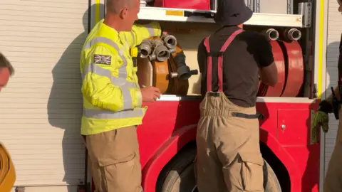 Four firefighters handling hoses at a fire engine on a sunny day.