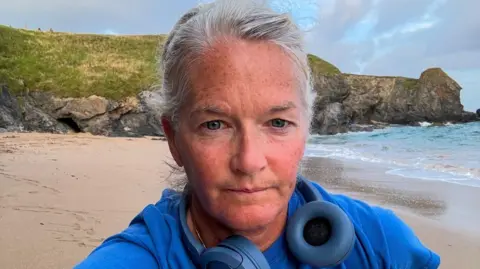 Rachel Clark. She has long, light grey hair which is tied back. A pair of blue headphones are around her neck and she is wearing a blue T-shirt. She has been running along a beach and the sand and sea can be seen in the background behind her. She is looking directly at the camera.