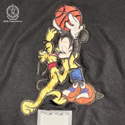 Polícia de Nova Gales do Sul Uma camiseta preta do Mickey Mouse com um buraco no meio e um cartão de provas colocado sobre ela.