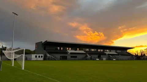 Cambridge City Football Club Cambridge City FC ground at Sawston. A grey football stadium stand and the above is a yellow/orange sunset. There is also a goal with the net folded up at the end of the pitch which runs in front of the stand