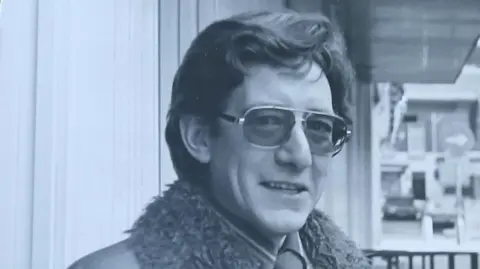 Supplied Black and white image of Gavin Petrie. He is wearing sunglasses and looking at the camera. In the background is a street scene. The fur top of his coat is visible.