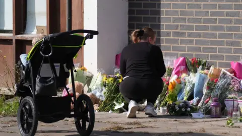 PA Media A woman crouches down in front of a number of bunches of flowers propped against a breeze-block wall. She is wearing black sportswear and white trained. A pushchair stands nearby.