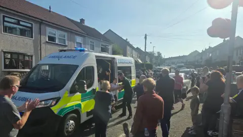 An ambulance on a residential street. Dáithí is being helped out by a paramedic. A crowd of people are standing on the street clapping.