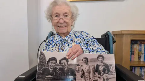 An elderly lady with white hair, glasses and a blue patterned dress smiling into the camera. In her hand is a photograph album showing images of her and her friends 80 years ago.
