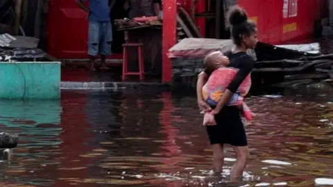 A girl carries an infant as she wades through water.