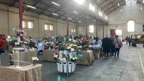 A view inside the market showing stalls selling flowers and other goods. The market has brick walls and a vaulted ceiling. 