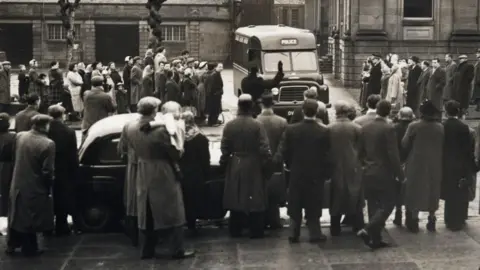 Getty Images A large crowd of onlookers stand outside the court building. A prison van is moving towards the crowd and a police officer can be seen holding his hand up in front of it. 