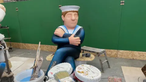 The foreground is covered with paint pots and brushes. In the background sits the figure of a sailor in blue uniform and white hat. He is missing all his facial features they have not been painted on yet.