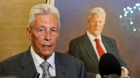 Peter Robinson is standing in front of some microphones. He is wearing a grey suit and a grey striped tie. He has white spiky hair and is face is very tanned. Behind him is a portrait of himself, which depicts him sitting down with grey hair and in a grey suit with a red tie.