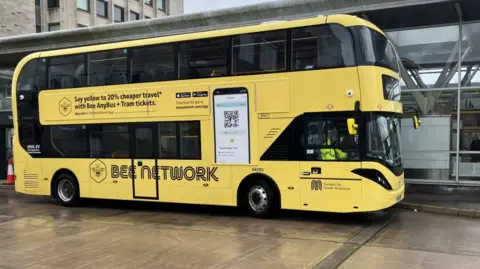 A yellow double-decker bus with the words 'bee network' on the side is pulled up beside a building. 