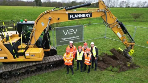 An aerial shot of six people wearing hi-vis jackets in a field, with a big yellow digger behind them.