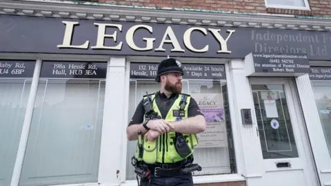 Danny Lawson/PA Media A male police officers stands outside a funeral parlour. He is wearing a black helmet and a yellow, high-vis vest over a black uniform. The funeral parlour has large glass windows and closed white blinds. A sign has gold letters on a black background, reading "Legacy". Smaller signs advertise the various branches of the business, with addresses around Hull.