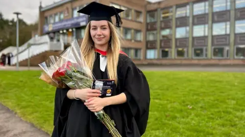 A woman with long blonde hair, wearing a black graduation outfit with a hat, smiles as she holds a bunch of flowers and a card. A university building is visible behind her.