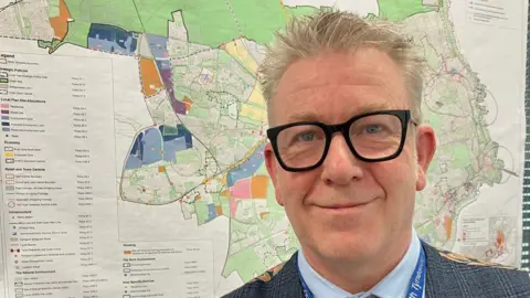 John Sparkes of North Tyneside Council. He is wearing dark-rimmed glasses and has grey hair. He is standing in front of a map of the proposed development site.