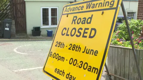 A yellow sign reading 'advance warning - road closed' stands to the side of a road in a small village