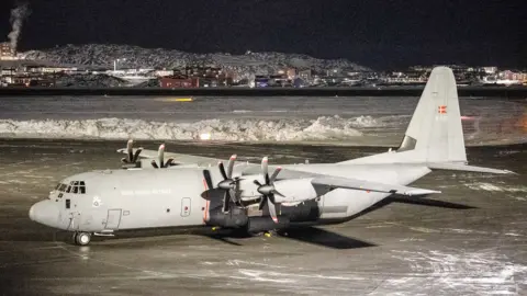 A Royal Danish Air Force Lockheed C-130J Super Hercules aircraft vehicle is parked on the tarmac at Nuuk international airport, Greenland. Snow has been cleared from the tarmac and is piled in an area behind the plane.. 