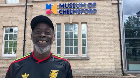 A man wearing a Jamaica football shirt and a cap stands outside a building with a sign that says "Museum of Chelmsford". He is smiling at the camera.