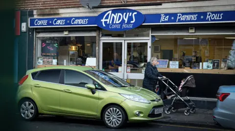 The exterior of Andy's Coffee House - a typical High street frontage with large windows and a blue and white sign, Cars are parked on the road outside and lights on in the shop make it look inviting.