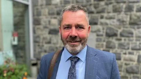 Deputy Marc Leadbeater. He is wearing a blue suit. He has short dark hair and a dark and grey beard. 