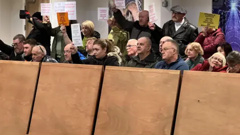 BBC Three rows of people are sitting behind some wooden boards at Carlisle Civic Centre's main council chamber. Many are holding signs with messages such as "Cumbria Action says no to council of sanctuary"