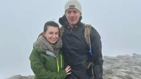 Family Emma and Jordan Fellows wearing walking kit are photographed at the top of a hill with the sky behind them.