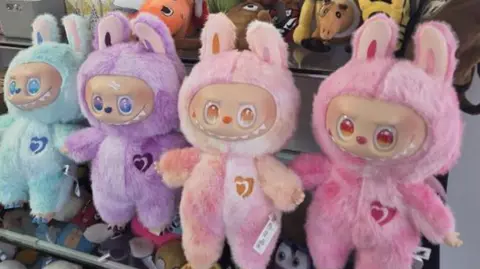 A row of  four furry and colourful fake Labubu dolls on a shelf. They have large eyes and jagged white teeth.