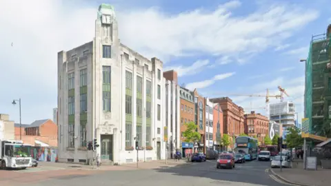 Google Maps Image of the former bank of Ireland in Belfast. It is a tall white art deco building with green detailing standing at a street corner in front of a junction. Traffic, including buses and cars, moves along the road beside rows of red‑brick and modern buildings under a bright, partly cloudy sky.
