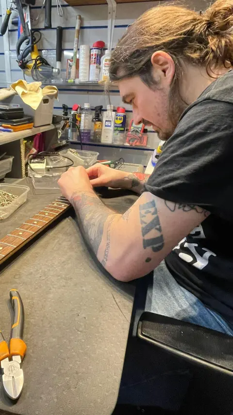 Mark Wood Sam Orr is working on a guitar neck, with tools around him.