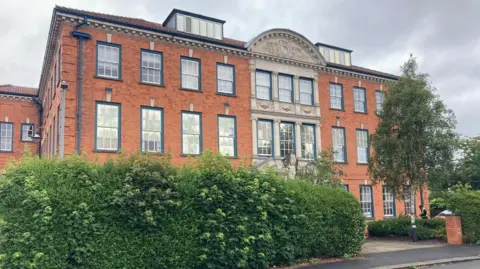 A three-storey building, seen mainly from the front, but with the image also featuring the left hand side of the building. Bushes are in front of the building.
