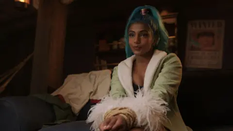 Netflix Chandran in character as Nefertari Vivi wears a green coat with fluffy sleeves and has blue hair tied up in a ponytail with a metallic headband