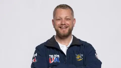 Royal British Legion Stephen Hooper, who is wearing a navy blue Invictus Games Team UK zip jacket. He has crossed his arms and is smiling at the camera.