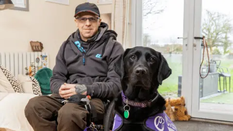 A man with glasses and a cap sitting on a bed wearing a blue top and brown trousers with a black dog sitting alongside him and a patio door behind them