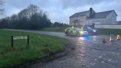 BBC Two police cars with their emergency lights on are blocking off the road. Three police officers can be seen wearing high vis yellow jackets. Yellow cones and a police warning sign are on the road in front of the cars. A street sign that says Killylea Road can also be seen.
