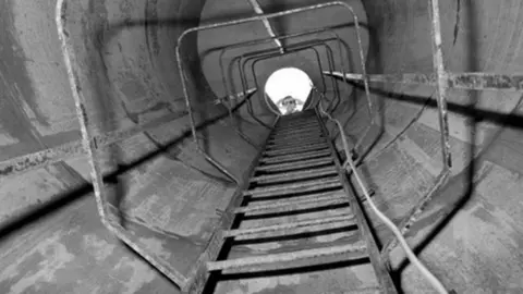 PA Media An old black and white picture from the bottom of a ladder in a drainage shaft, looking up towards the surface. A light-coloured wire is seen dangling down on the right. A person can be seen looking down from the top of the ladder.