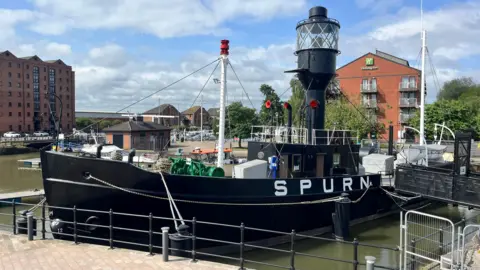 The picture shows a large black ship which has been docked. It reads 'SPURN' in white block capitals on the right hand side. 