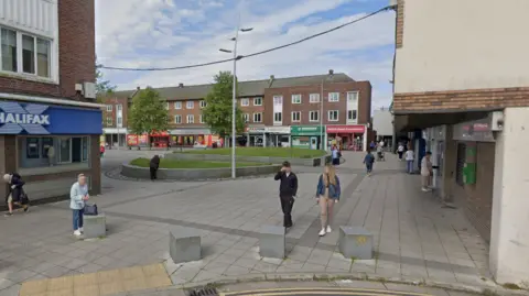 A courtyard in Billingham town centre has some greenery in amongst the concrete slab floor. Shops surround the square and people are walking through.