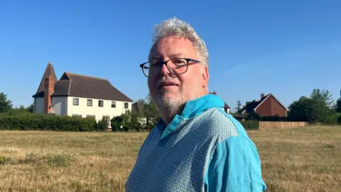 Holly Nichols/BBC Brian Cheyne standing in a field with two detached houses behind him. He has white hair and a very short white beard. He is wearing black-framed glasses and a bright blue shirt.