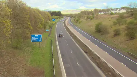 M90 with cars on it. There are tyres and houses surrounding it and the sun is shining on hills in the distance.