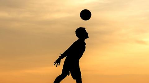 A young player heads a football at sunset
