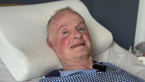 Brian Shields, a man with grey hair, wearing a checked blue top, with his head on a pillow.
