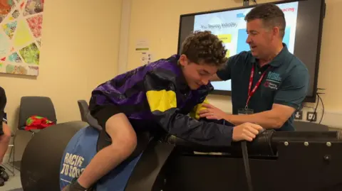A young boy on a metal race horse machine, with a man showing him, how it works. They are in a classroom setting.
