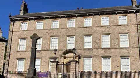 Exterior shot of Lancaster Judges Lodgings. It is an early 17th century townhouse, made of stone, with three sash windows either side of the front door and seven windows on the first and second floors. There are iron railings in front of the building and there is a stone cross to the left of the front gates.
