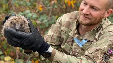 Sgt Paul Smith, on the right, in camouflage uniform, wearing black gloves and holding a hedgehog who is facing the camera.