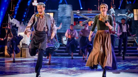 Carlos Gu and Karen Carney, both wearing old-fashioned clothes, smile widely as they dance on the Strictly Come Dancing dancefloor. More dancers can be seen behind them.