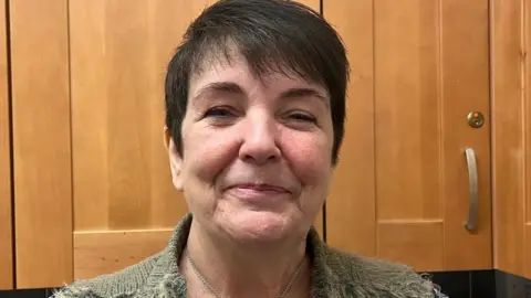 A woman with short dark hair looks straight into the camera with a hint of a smile. She is in front of wooden kitchen cupboards and the shoulders of a woolley grey jacket can be seen.